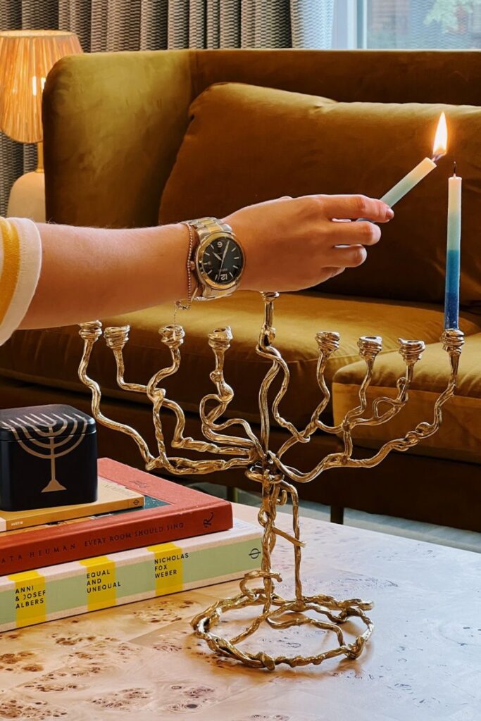 Image of a woman's hand extending to light a menorah candle, she is wearing a large watch and dainty tennis bracelet.